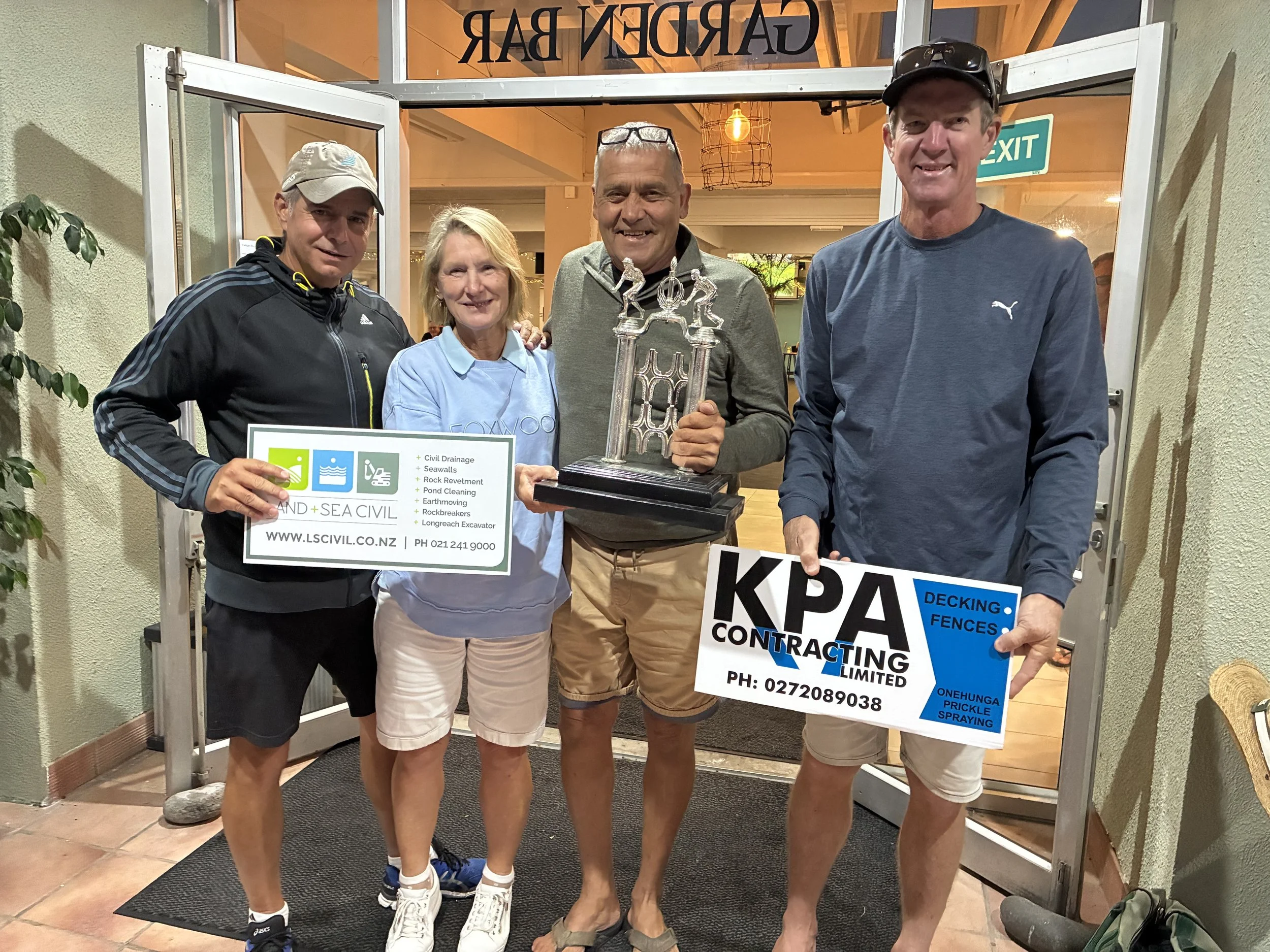 Four people standing together outside a building, holding signs and a trophy, smiling for the camera. The group includes two men, one woman, and one person partially obscured. The signs advertise land and sea civil services and fencing contracting. T