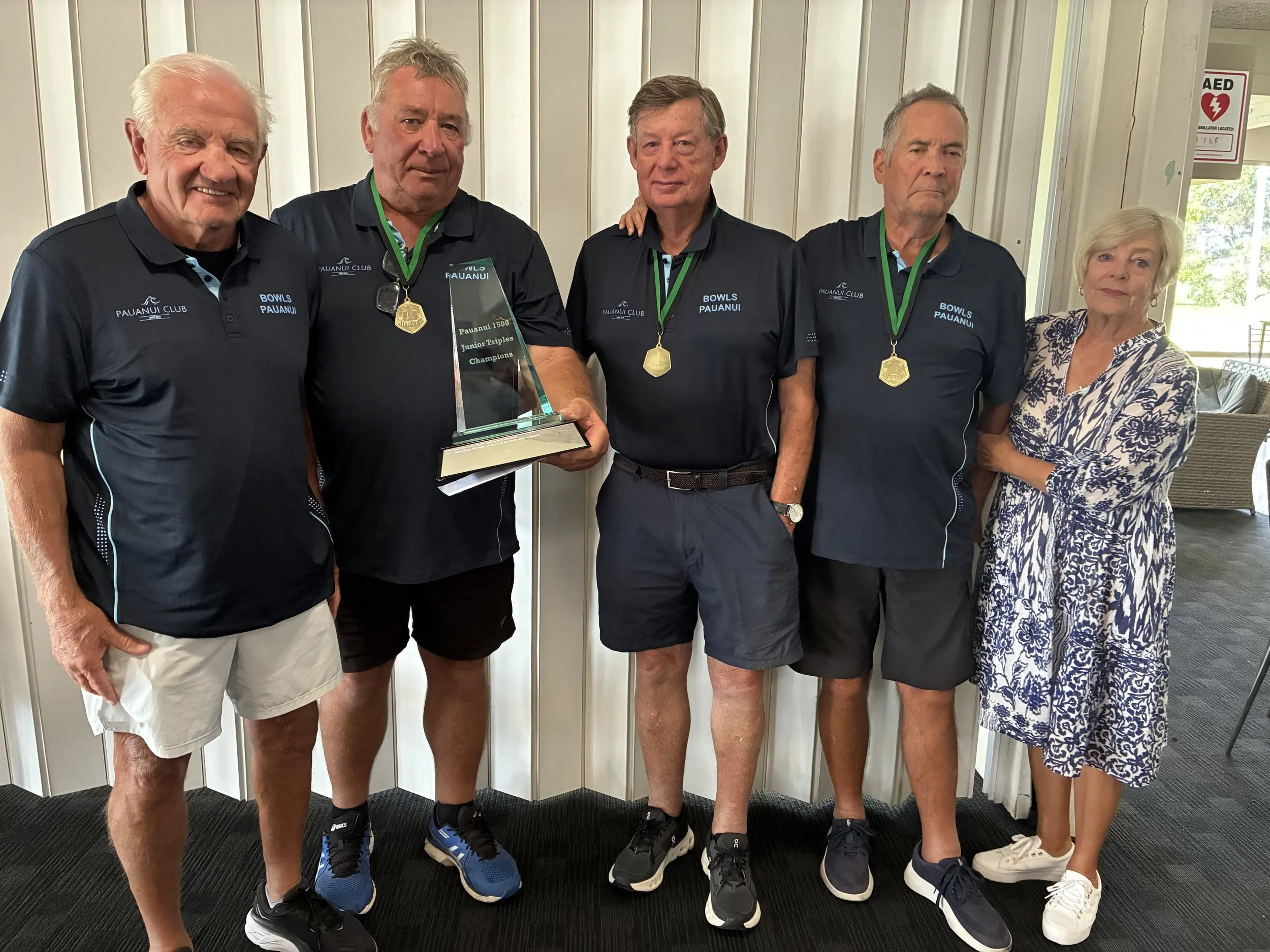 Four men and one woman standing together indoors, with the three men in black shirts with blue accents, medals around their necks, and one holding a trophy, all posing for a photo.