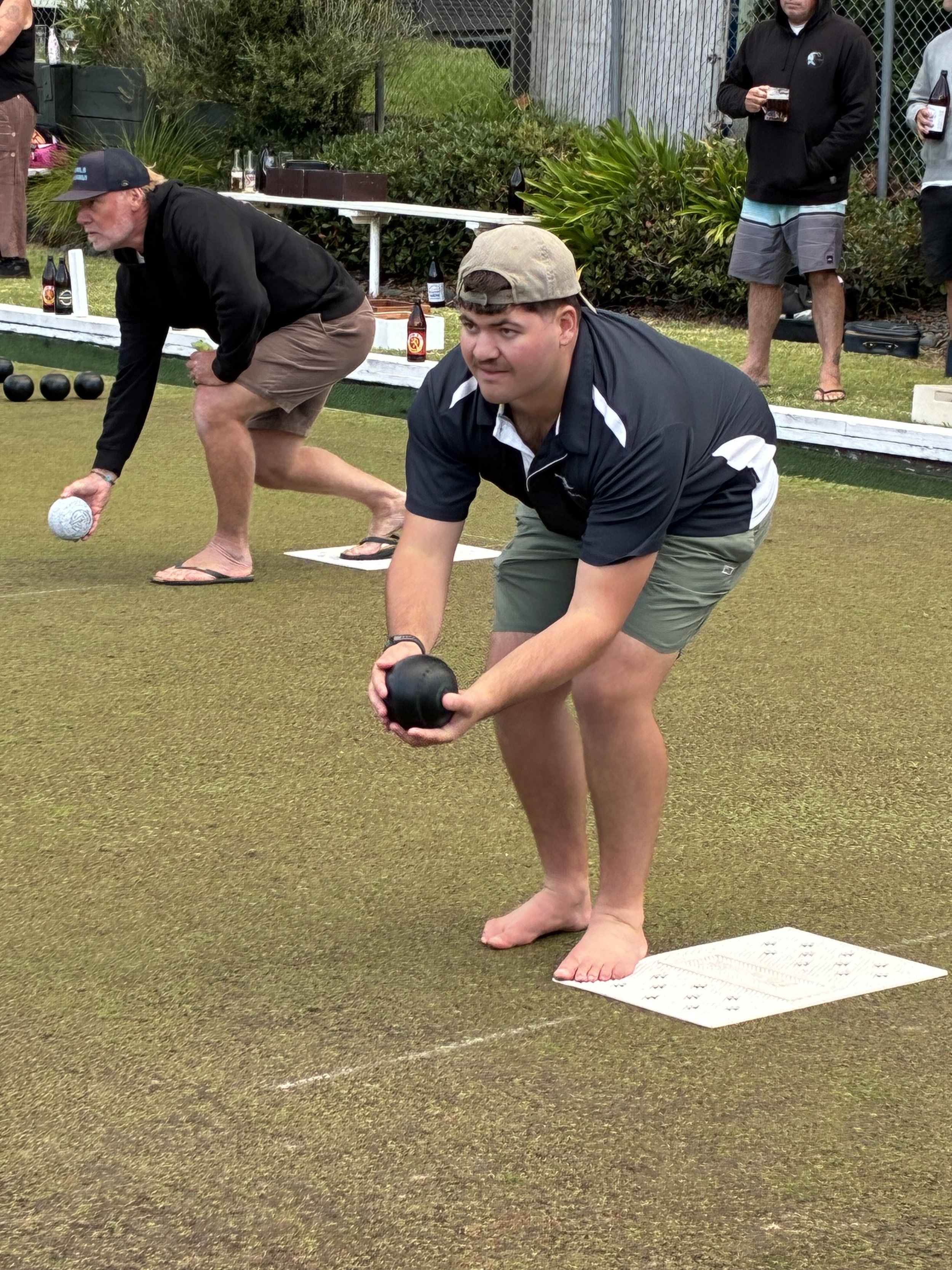 Two men are playing bocce on a grassy court, with several bottles of beer on a table in the background. One man in the foreground is wearing a black jacket, khaki shorts, and a backwards cap, and is holding a bocce ball. The other man in the middle g