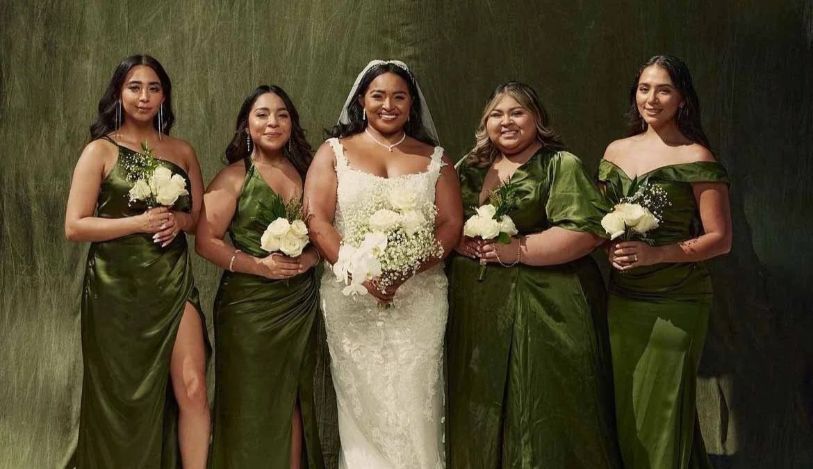 A bride in a white wedding dress and four bridesmaids in matching dark green dresses, standing together and holding white bouquets, in front of a dark textured green background.