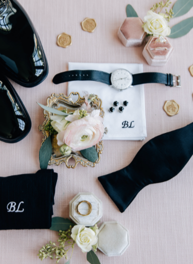 Flat lay arrangement of accessories and decor on a pink surface, including black shoes, a watch, wedding rings in a hexagonal box, a bouquet of white and pink flowers, earrings, monogrammed cloths, and small decorative objects.
