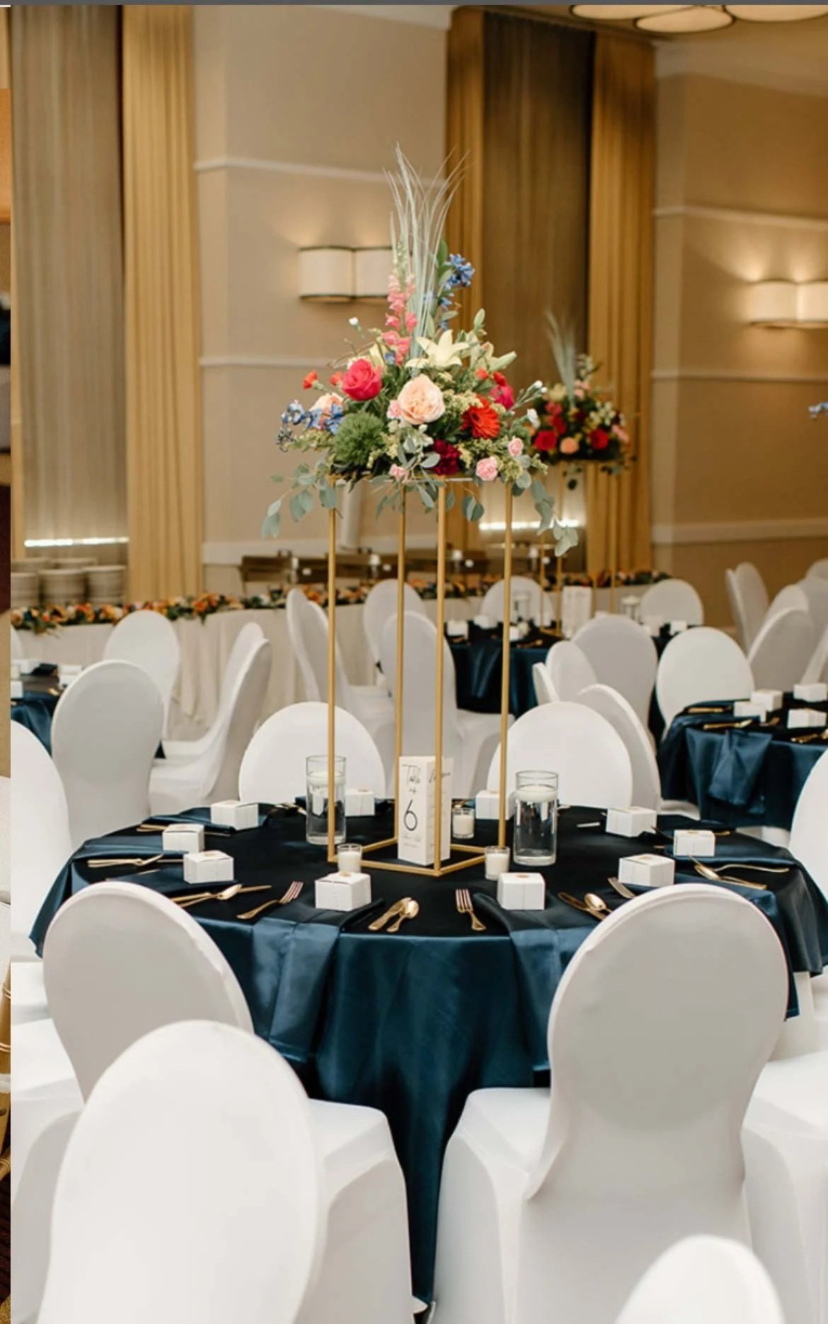 Elegant banquet table decorated with a tall floral centerpiece of pink, red, and white flowers. The table is set with black tablecloths, gold cutlery, white square plates, water glasses, small candles, and small white boxes. The chairs are white, and