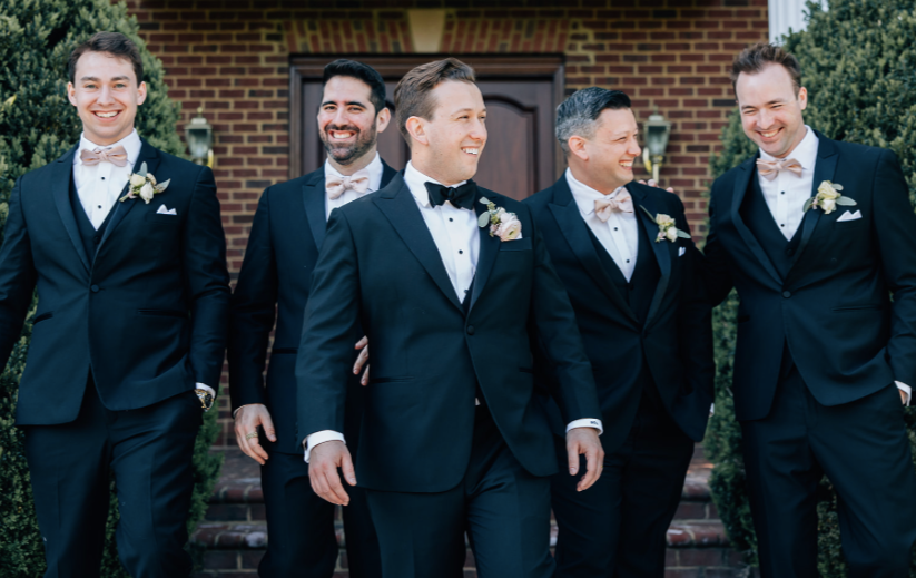 Five men in tuxedos walking together outside in front of a brick building, smiling and laughing.
