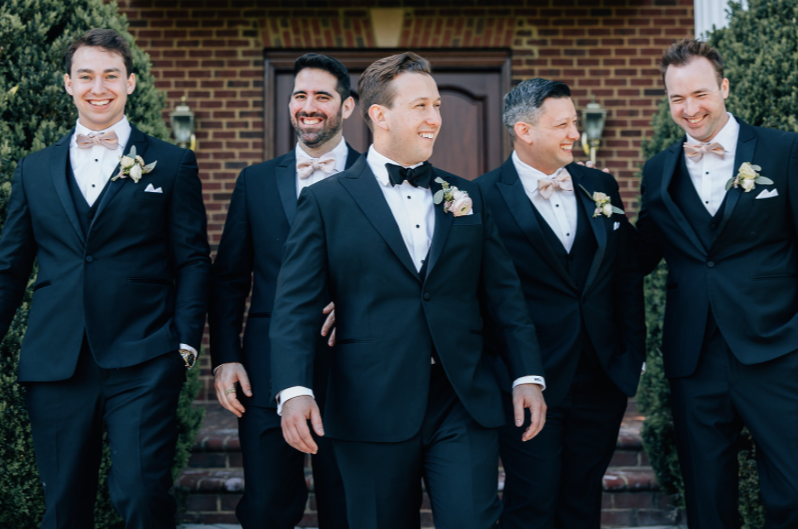 Group of five men in tuxedos walking outdoors in front of a brick building, smiling and enjoying each other's company.