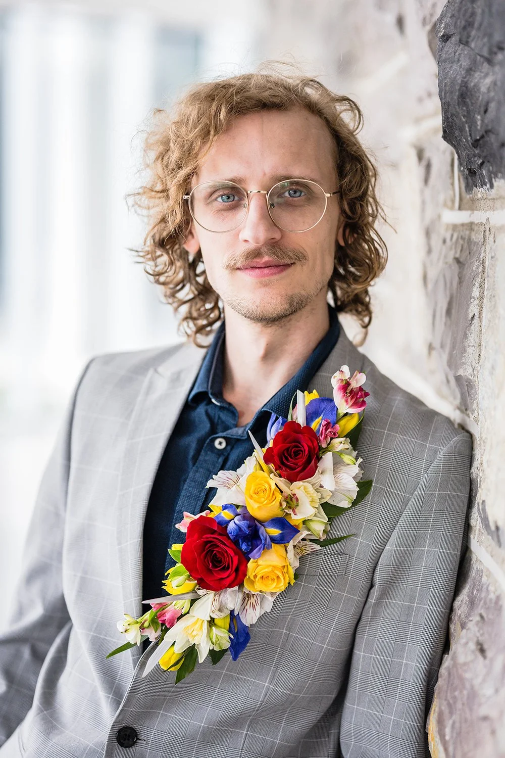 Man in a gray suit posing against a wall wearing a navy blue shirt, and a floral lapel boutonniere.