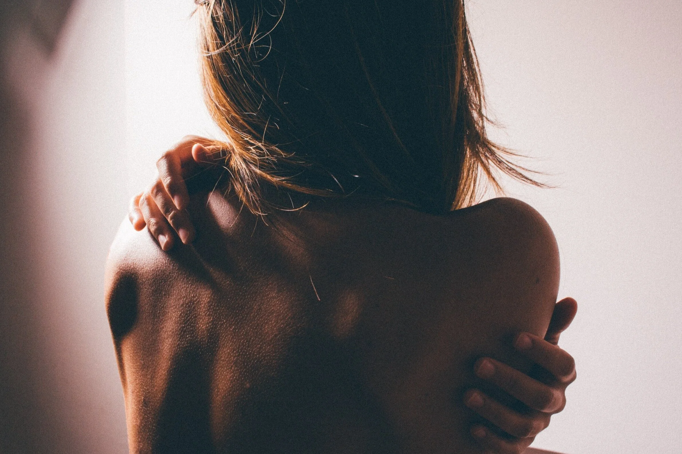 A woman with shoulder-length hair is touching her shoulder and back, with her face turned away from the camera, in natural lighting.