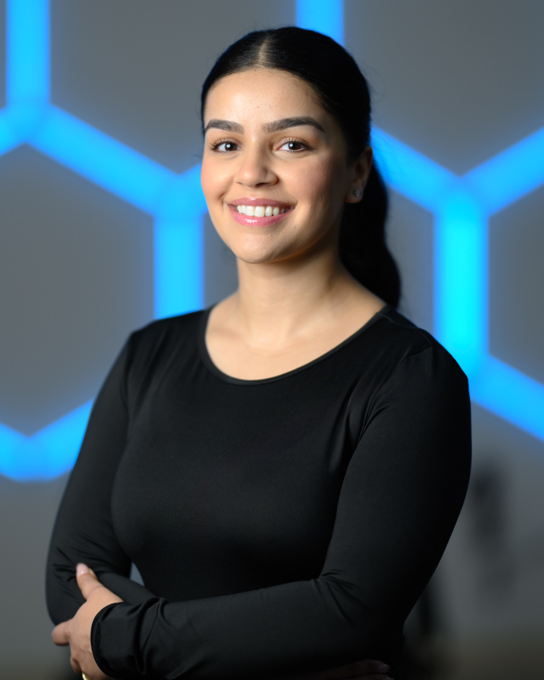 A young woman with long black hair and a bright smile, standing against a plain white wall.