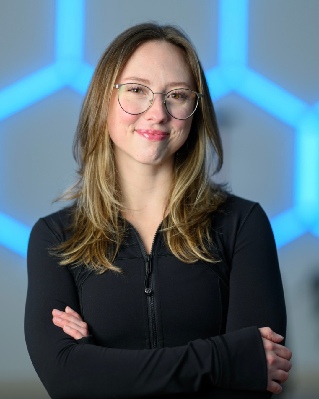 A young woman with long, wavy light brown hair wearing round glasses, smiling at the camera.