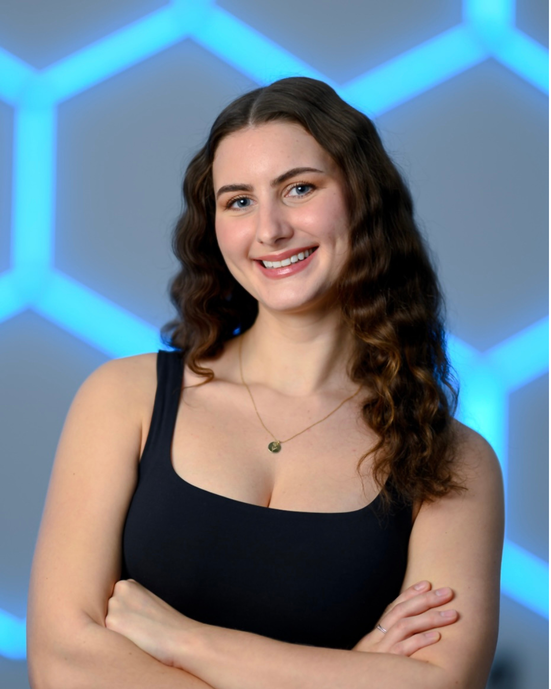 A young woman with long wavy brown hair, blue eyes, and light skin, smiling while standing against a plain, light-colored background. She is wearing a sleeveless, fitted blue top.