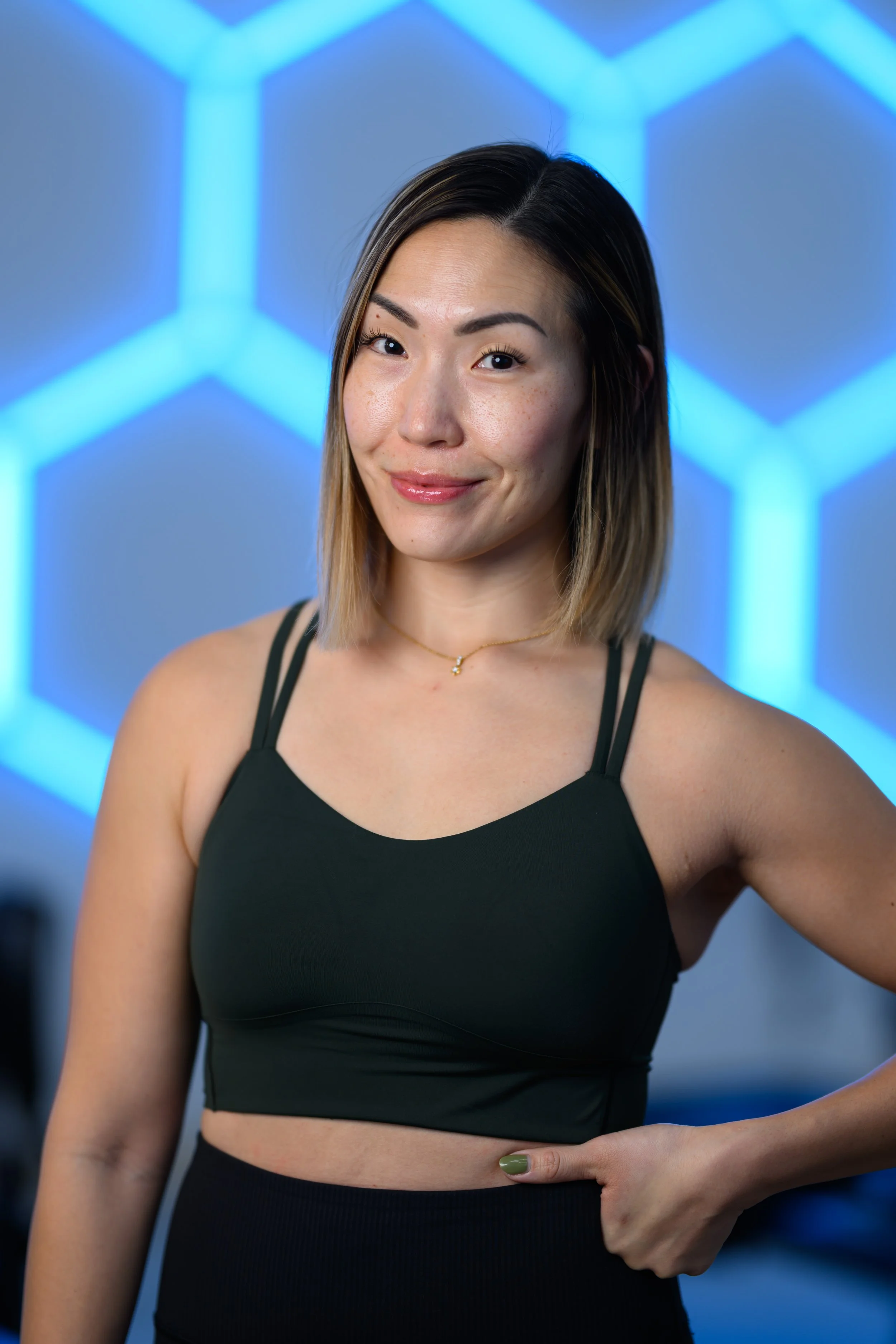 A woman with shoulder-length light brown hair, wearing a blue sleeveless top and a delicate necklace, smiling at the camera against a plain, light-colored background.