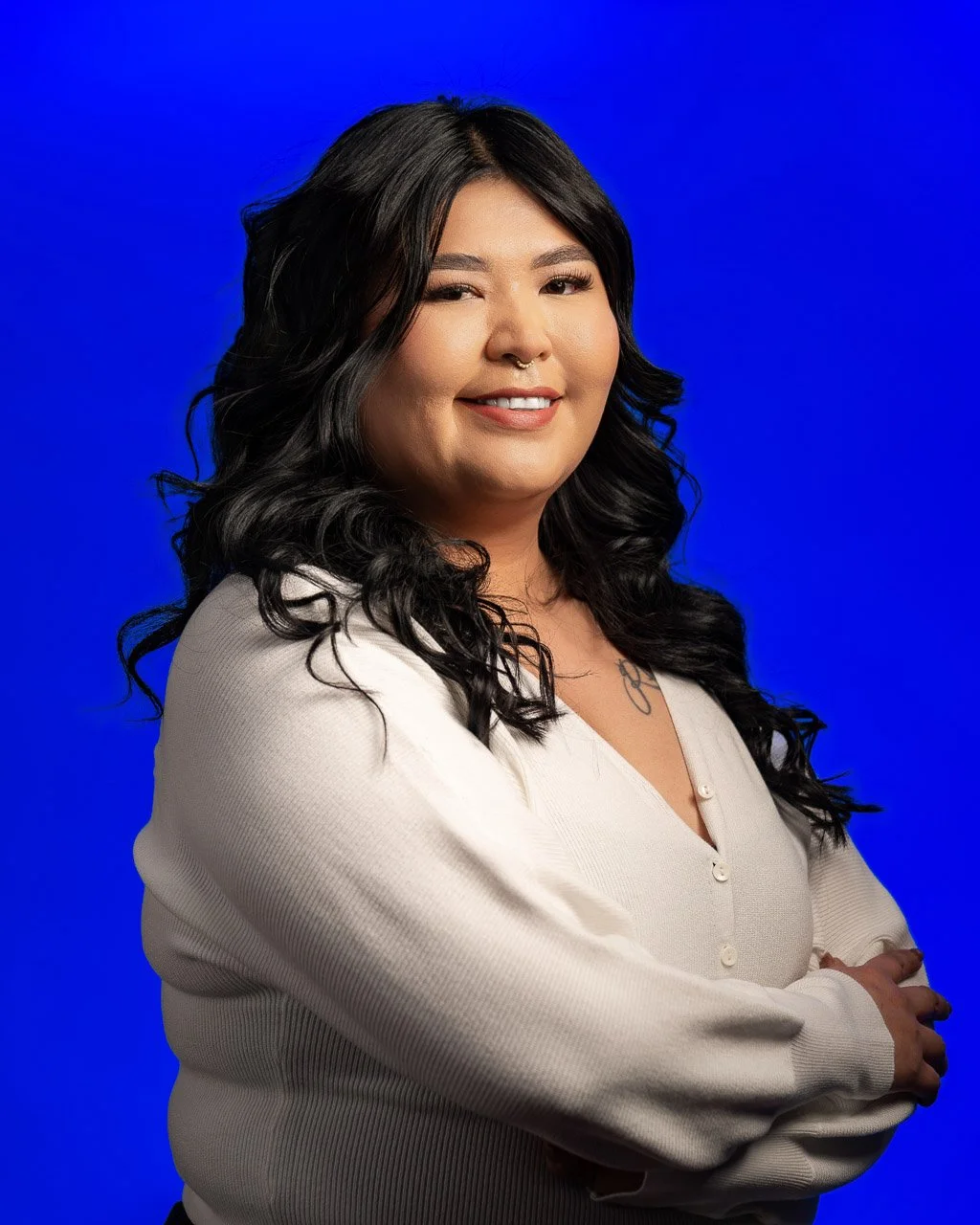 A woman with long black wavy hair, wearing a light-colored blouse, standing against a solid blue background, smiling with arms crossed.