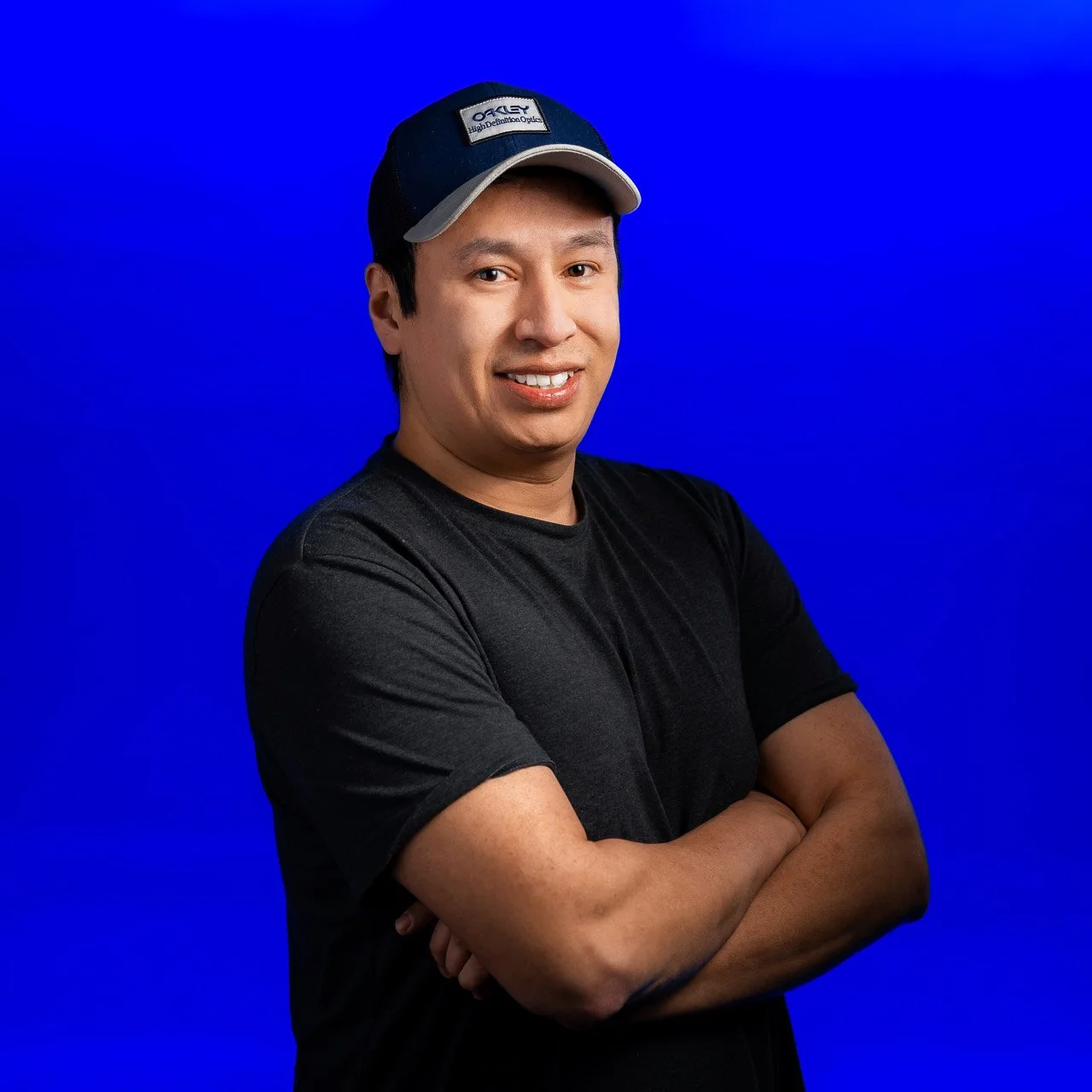A man in a black t-shirt and a baseball cap with a logo, standing with arms crossed in front of a solid blue background.