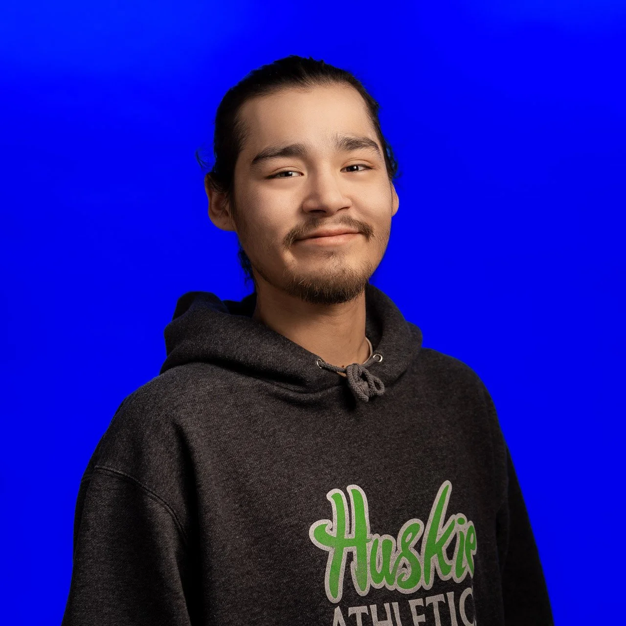 Portrait of a young man with long dark hair and a goatee, wearing a black hoodie with green and white text, smiling slightly, against a solid blue background.