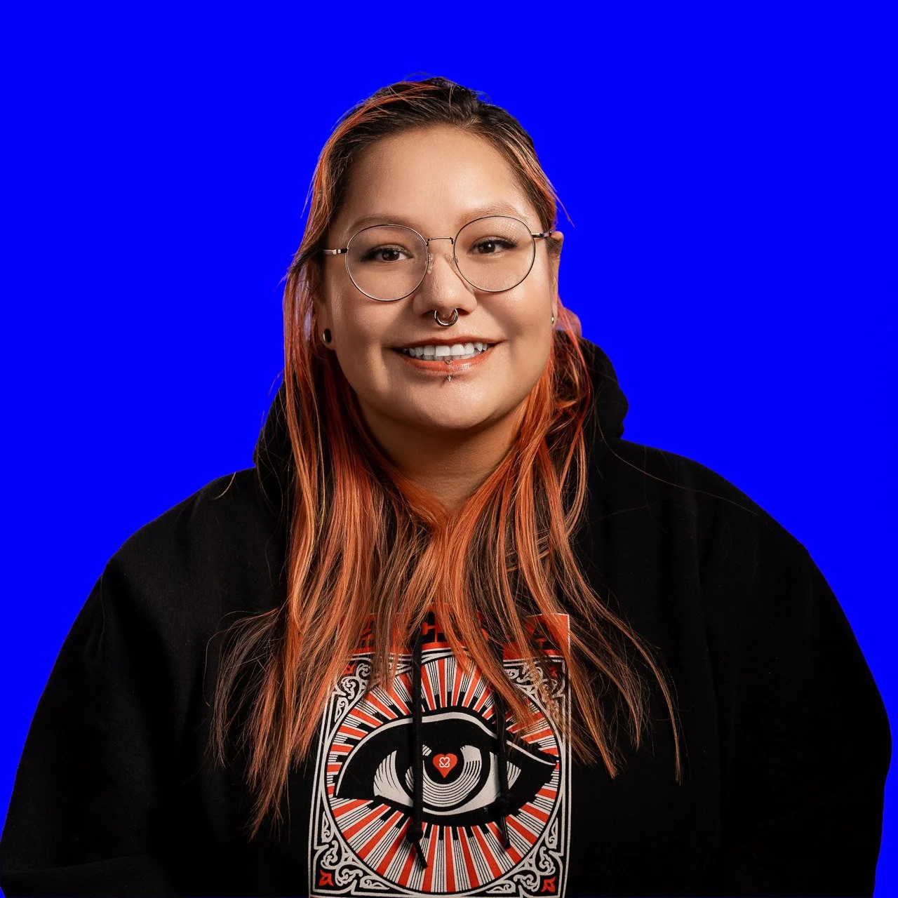 A woman with long reddish hair, glasses, and multiple facial piercings smiling in front of a solid blue background, wearing a black hoodie with a graphic design on the front.