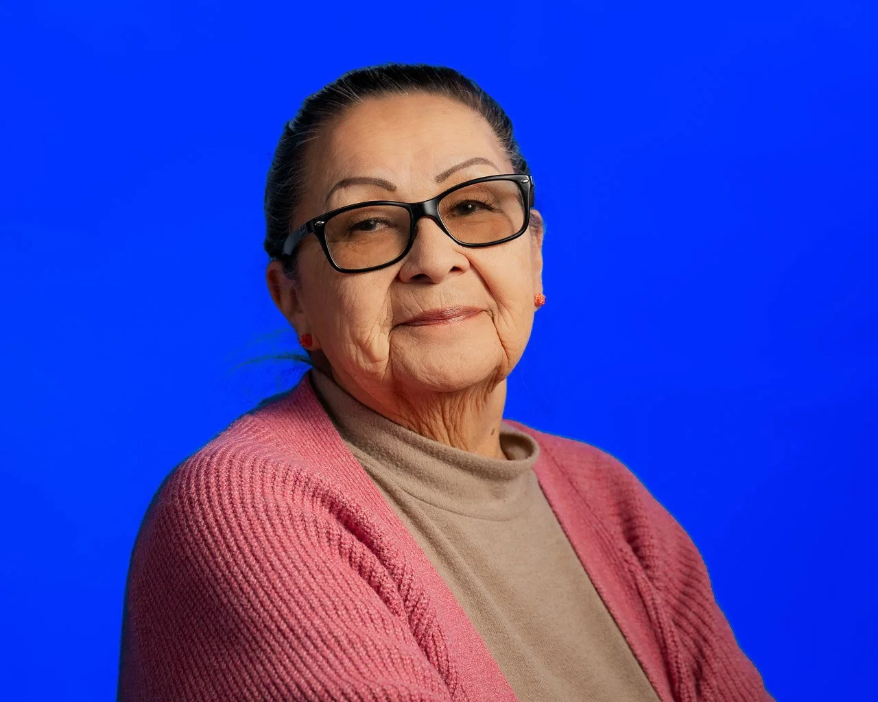 An elderly woman wearing glasses, a beige turtleneck, and an orange collared shirt, smiling.