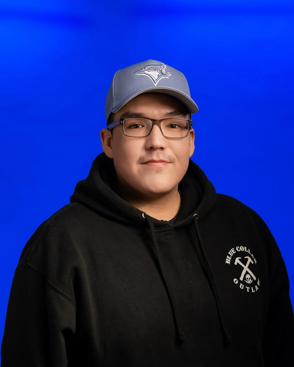 A young man with glasses wearing a gray baseball cap and a black hoodie with logo, posing against a solid blue background.