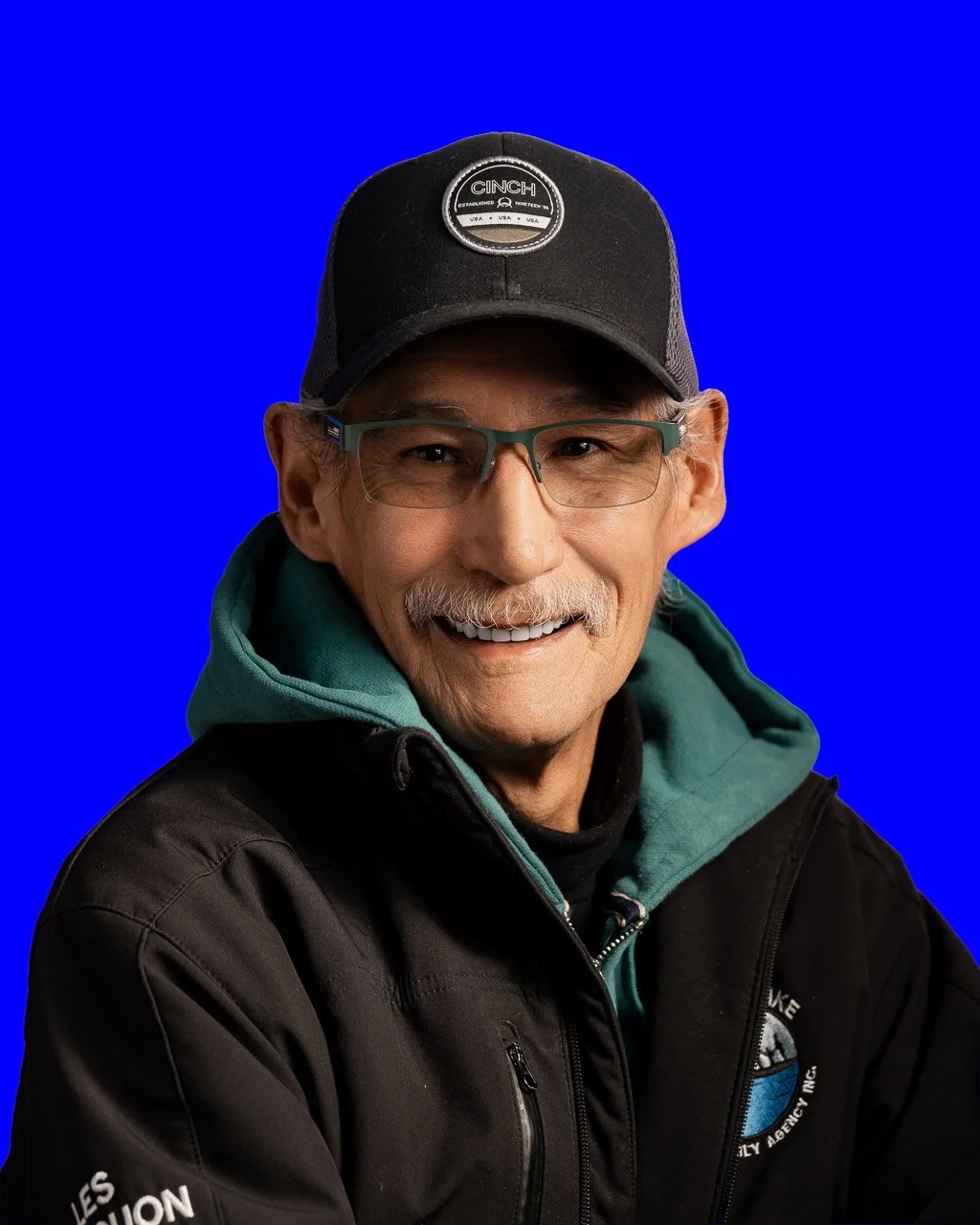 Portrait of a smiling elderly man with glasses, a mustache, wearing a black snapback cap with the word 'CINCH', a teal hoodie, and a black jacket with a logo, against a plain blue background.