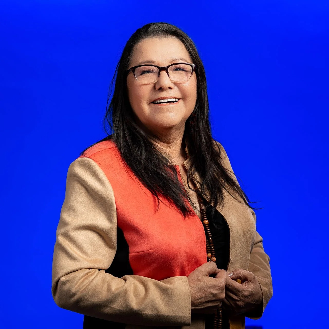 A woman with long dark hair and glasses smiling, wearing a color-blocked jacket in beige, red, black, and tan, standing against a blue background.