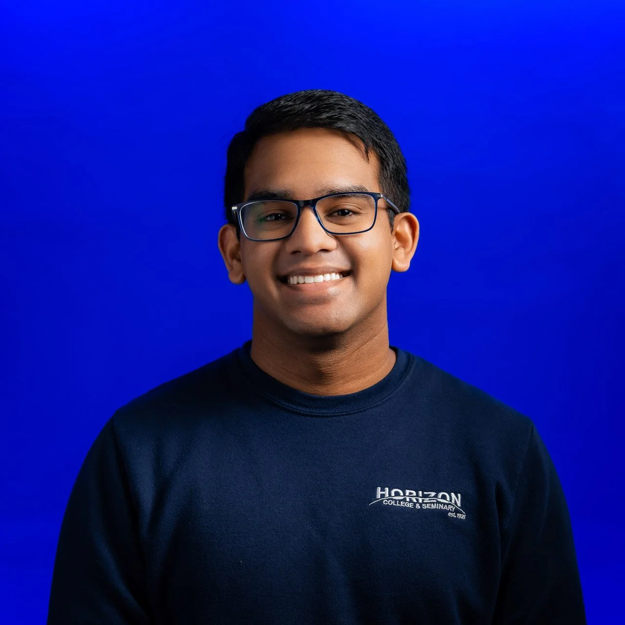 Portrait of a young man with short black hair, glasses, smiling, wearing a dark sweatshirt with 'Horizon College & Seminary' logo, against a bright blue background.