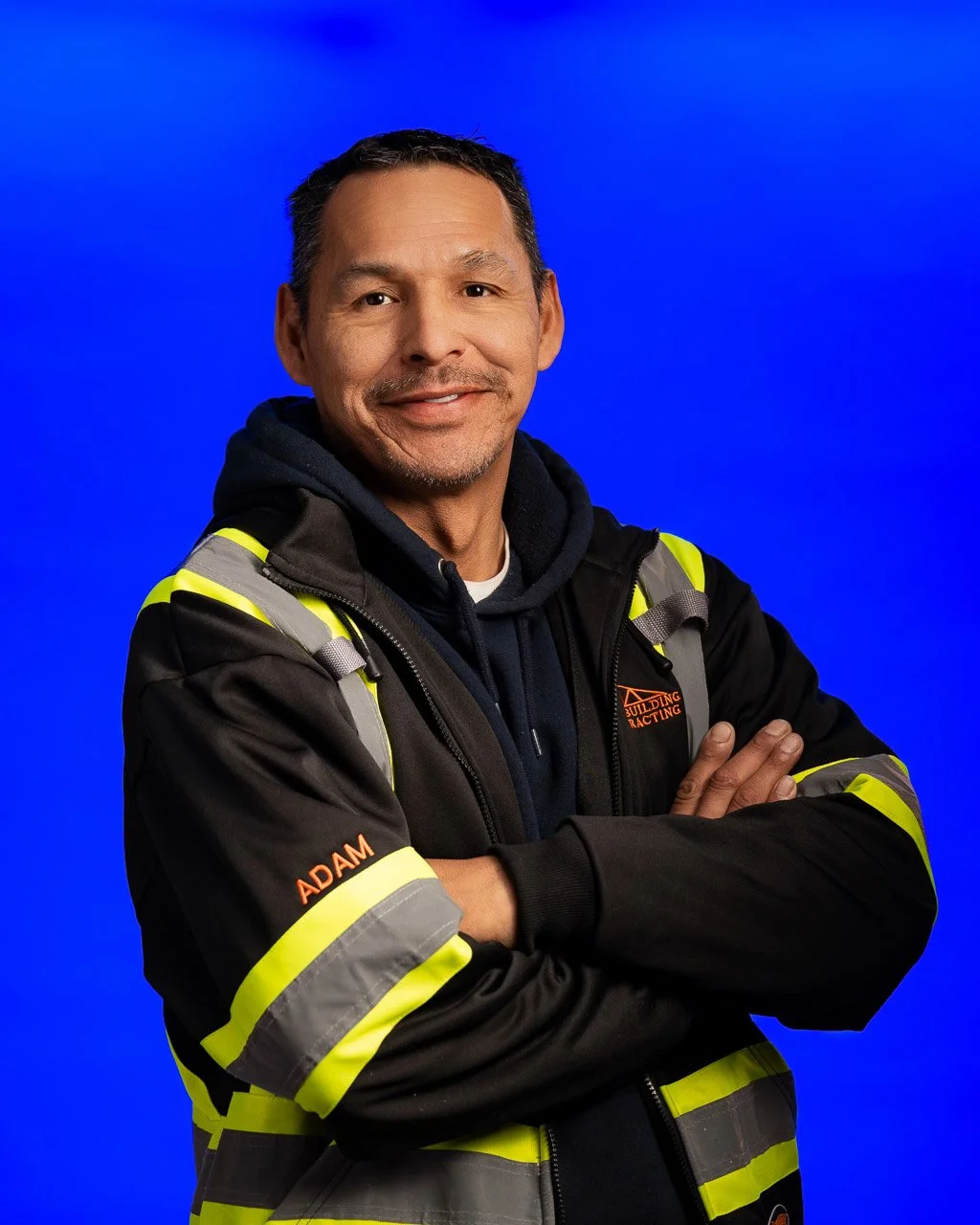 A man wearing a black safety jacket with neon yellow reflective stripes and the name 'Adam' embroidered on the sleeve, standing with arms crossed against a blue background.