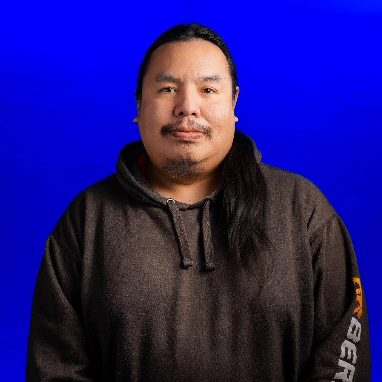 Portrait of a man with long black hair, facial hair, wearing a dark brown hoodie, standing against a vibrant blue background.