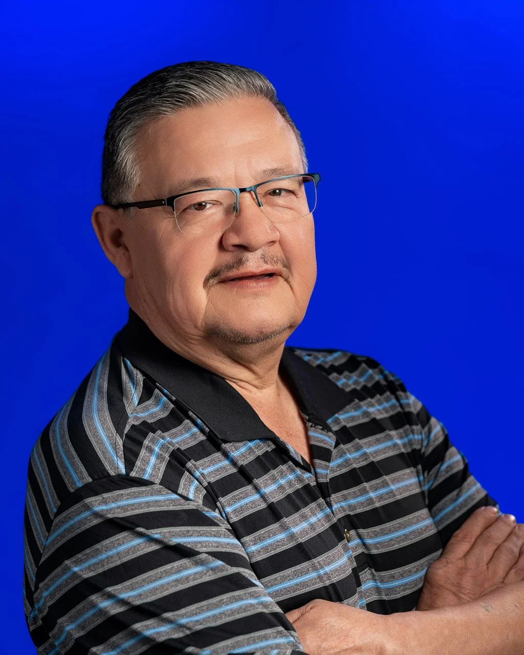 A middle-aged man with gray hair, glasses, and a goatee, wearing a black, gray, and blue striped polo shirt, standing with his arms crossed against a solid blue background.