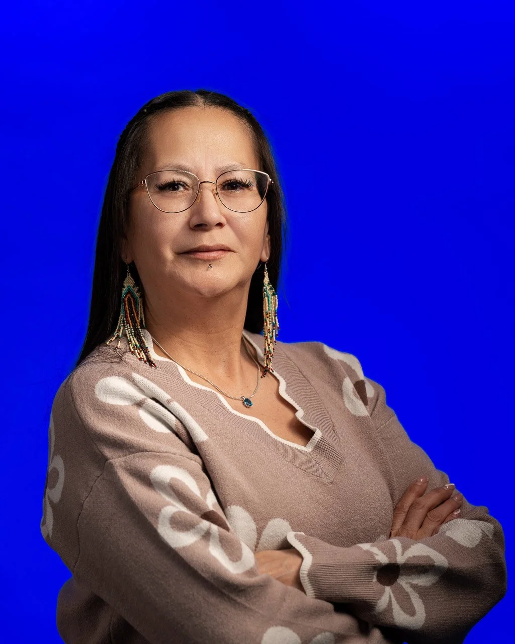 Portrait of a woman with glasses and beaded earrings standing against a plain blue background.