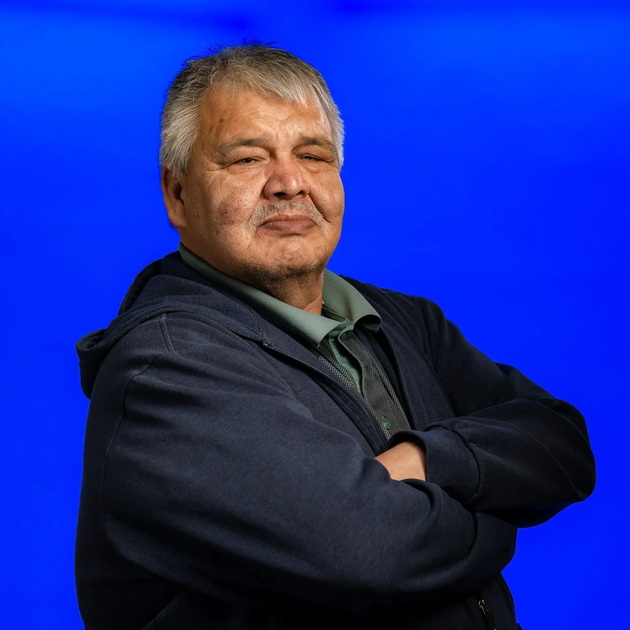 An older man with gray hair and a mustache standing with arms crossed in front of a blue background.