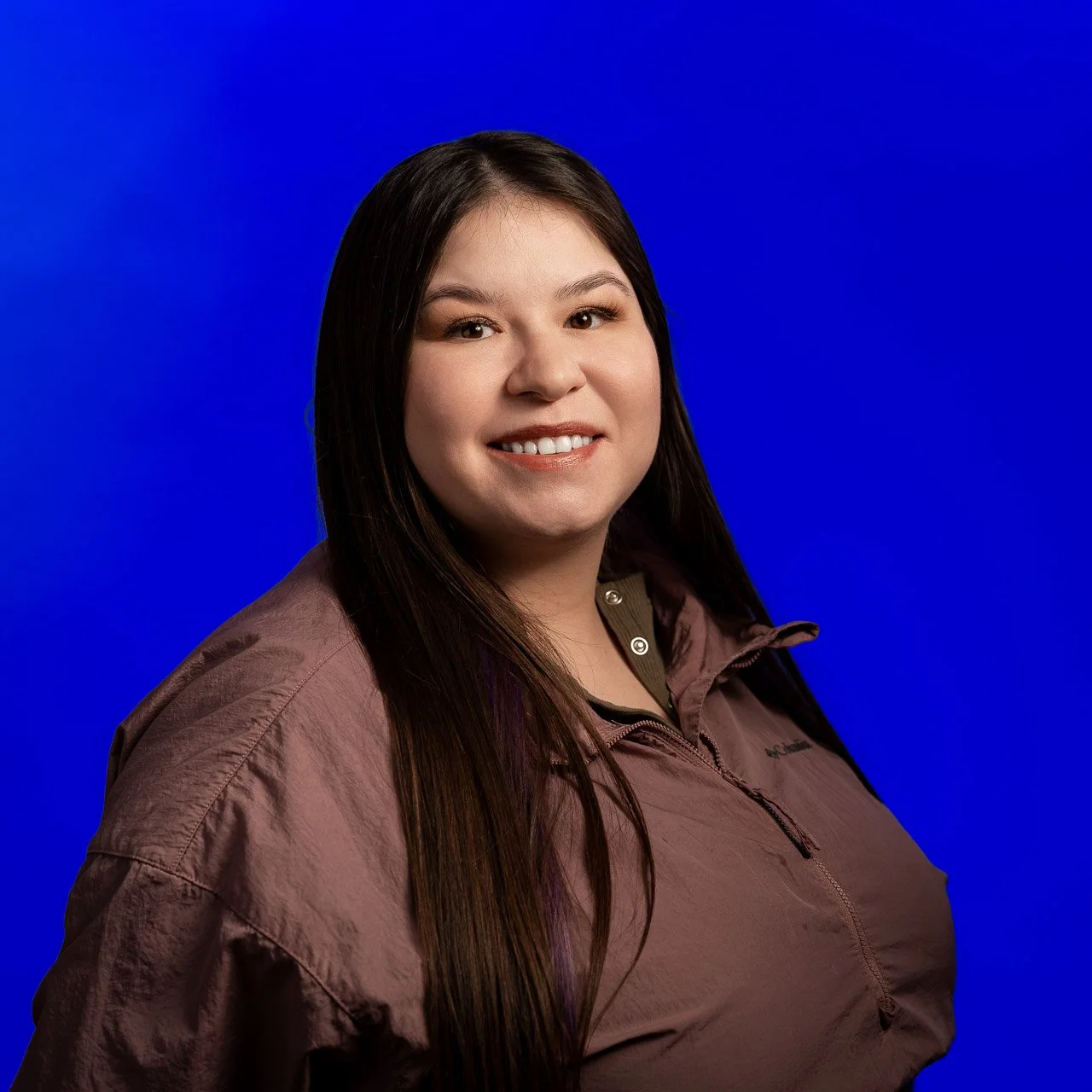 Portrait of a smiling woman with long dark hair, wearing a brown jacket, against a solid blue background.
