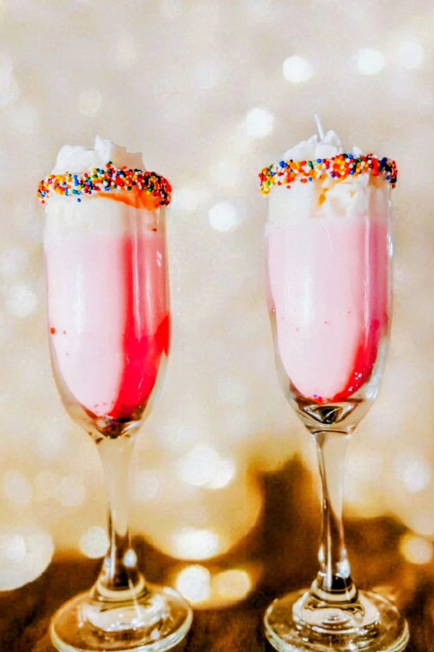 Two glasses of pink drink candles topped with wax whipped cream, sprinkles, and sparkly background.