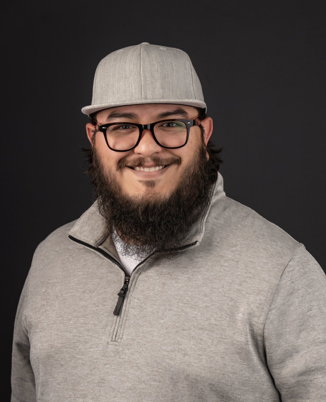 A man with a beard, glasses, wearing a gray cap and a gray zip-up jacket, smiling in front of a dark background.