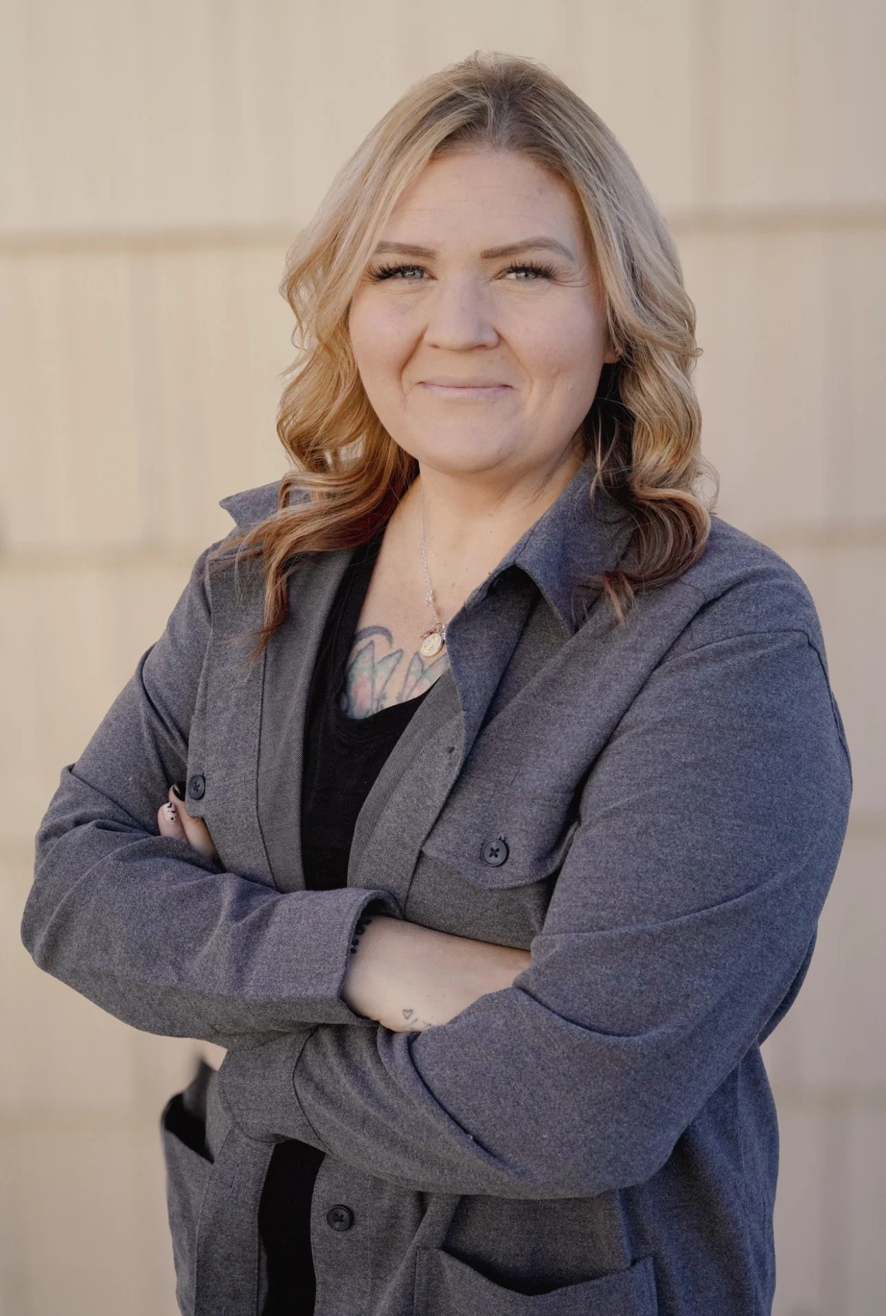 A woman with blonde, wavy hair crossing her arms, wearing a gray blazer and black top, smiling slightly in front of a light beige wall, with tattoos visible on her chest and wrist.