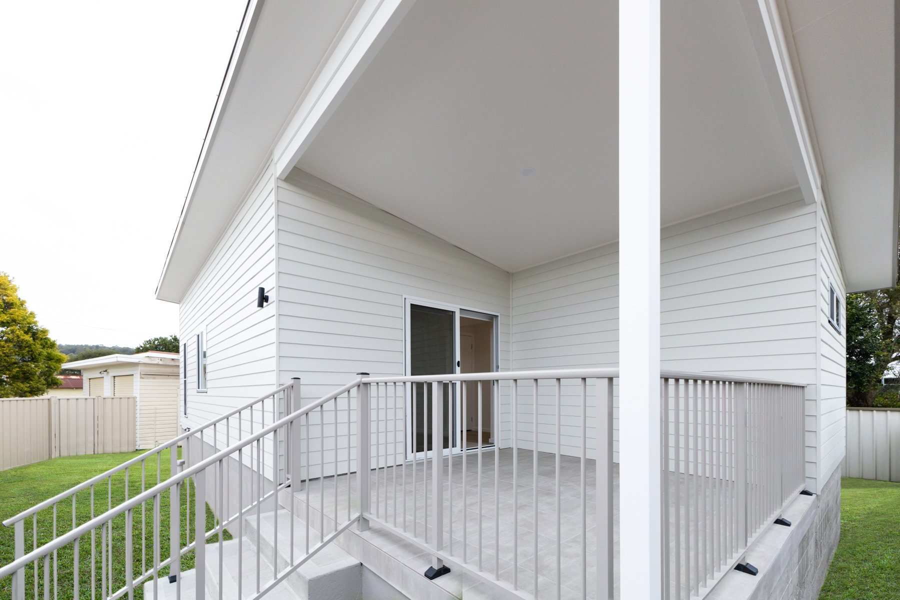Front porch of a white granny flat with a doormat, sliding glass door, metal railing, and green lawn