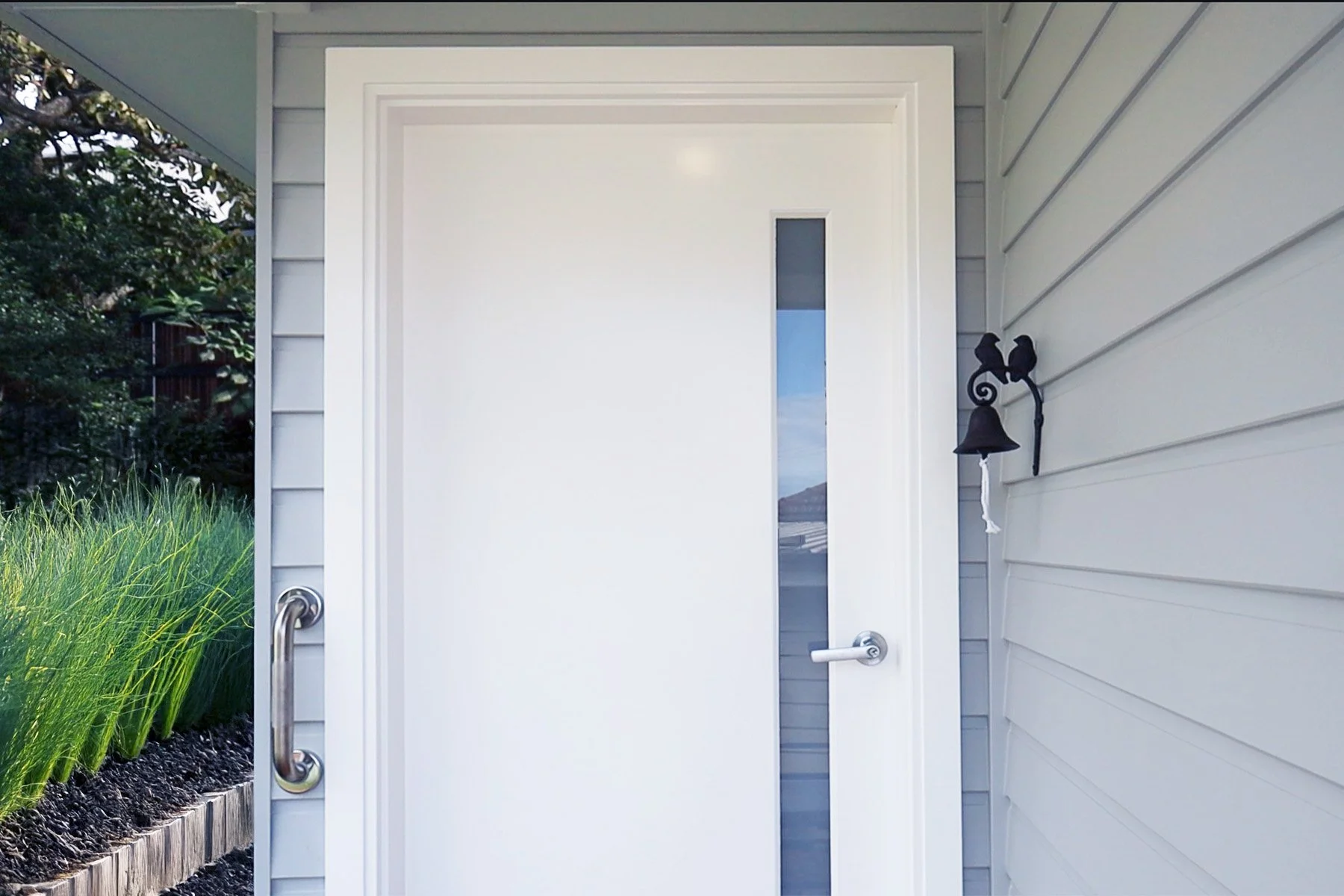 White front door with a vertical window pane, next to a black bell and light fixture on the beige siding of a house, with green plants on the left.