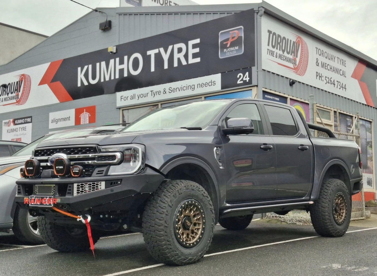 A black pickup truck parked outside a tire shop called Kumho Tyre, with a rugged off-road appearance, large tires, and aftermarket modifications including a front bumper and auxiliary lights.