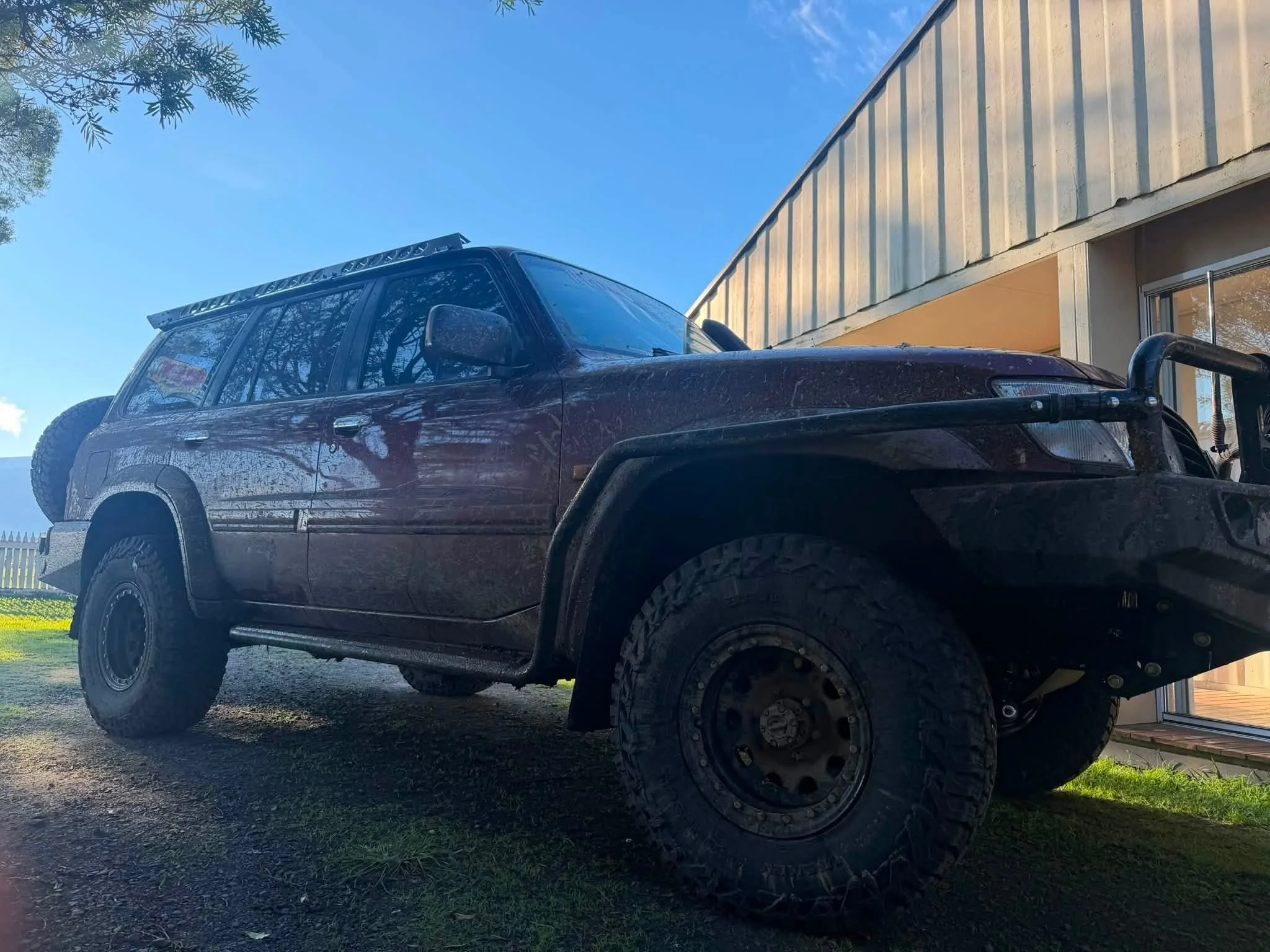 A black off-road vehicle parked on a driveway in front of a house with a metal wall exterior, under a clear blue sky, with trees visible in the background.