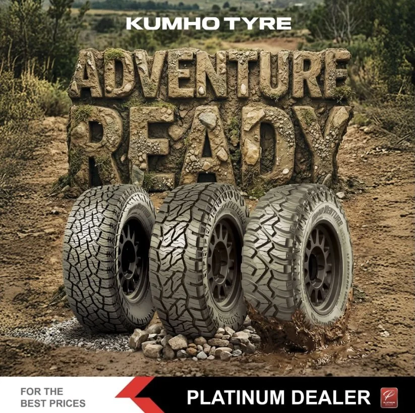 Three Kumho tires in front of a rocky landscape with a sign that reads 'Adventure Ready,' promoting a platinum dealer.