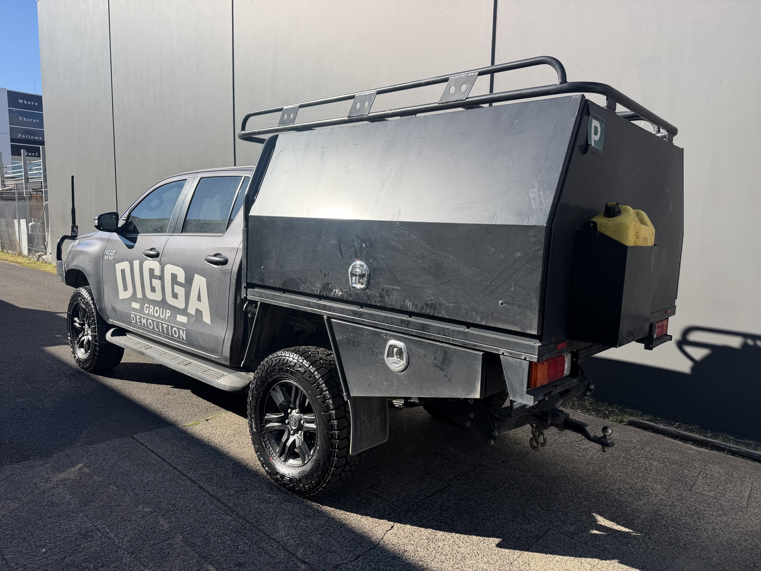 A silver pickup truck with branding for DIGGA Group Demolition parked next to a large beige wall. The truck has a black storage compartment on its back and a yellow container attached on the side. The vehicle is on a paved surface with some shadows and a clear blue sky is visible.