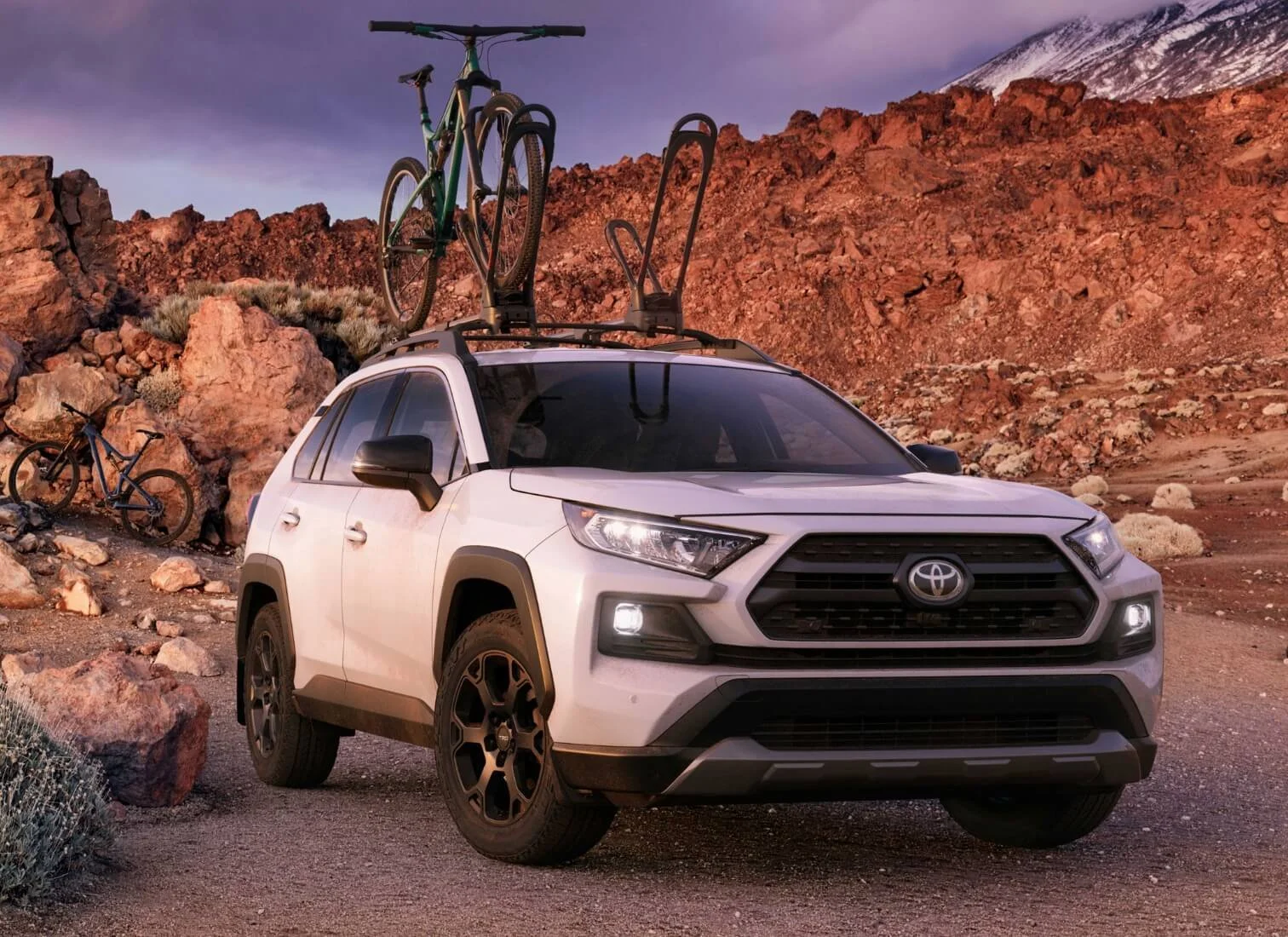 White Toyota SUV with bikes mounted on roof rack parked in a rocky, desert landscape with red rocks and mountain in the background.