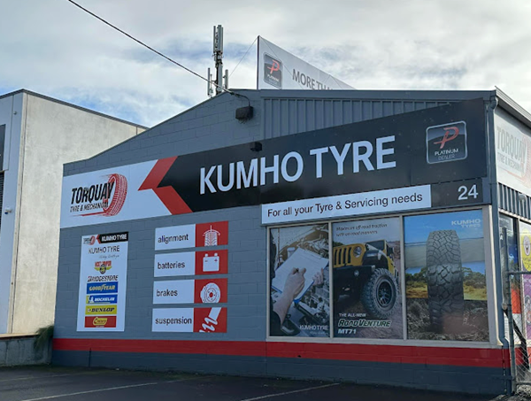 Exterior of a building with signage for Kumho Tyre, displaying services like alignment, batteries, brakes, and suspension, with images of off-road vehicles and tire services.