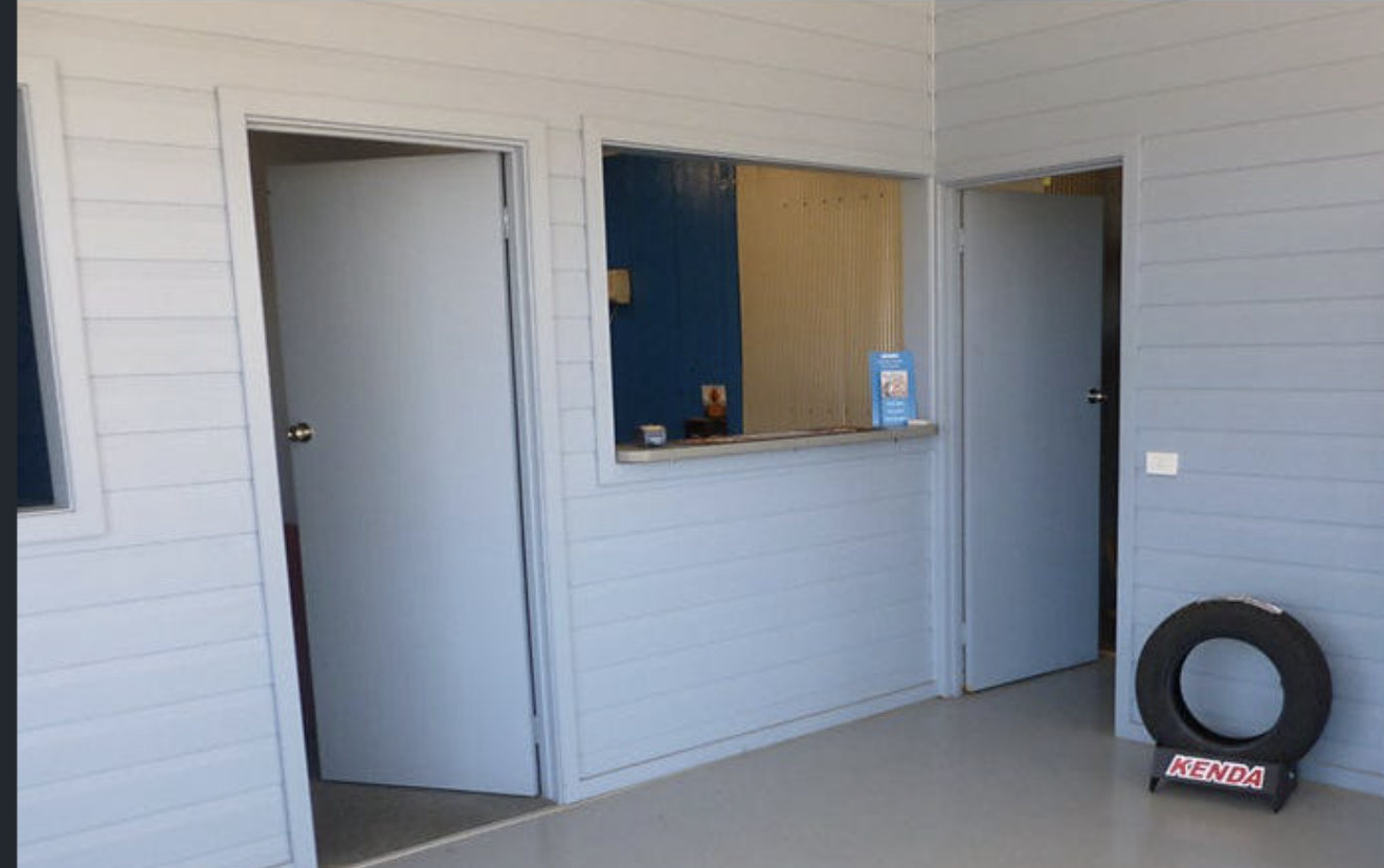 Interior of a building with white paneled walls and two open doors. There's a tire with a Kenda label resting against the wall. A service window with a small ledge separates this room from another room with blue and yellow paneled walls.