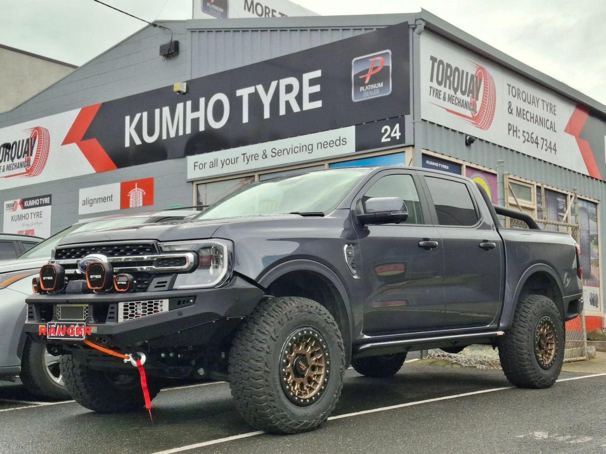 A black off-road vehicle with large tires, a winch, and orange tow strap parked in front of a tire shop, Kumho Tyre, with signage for Torque Tyre & Mechanical and advertising for platinum dealer services.