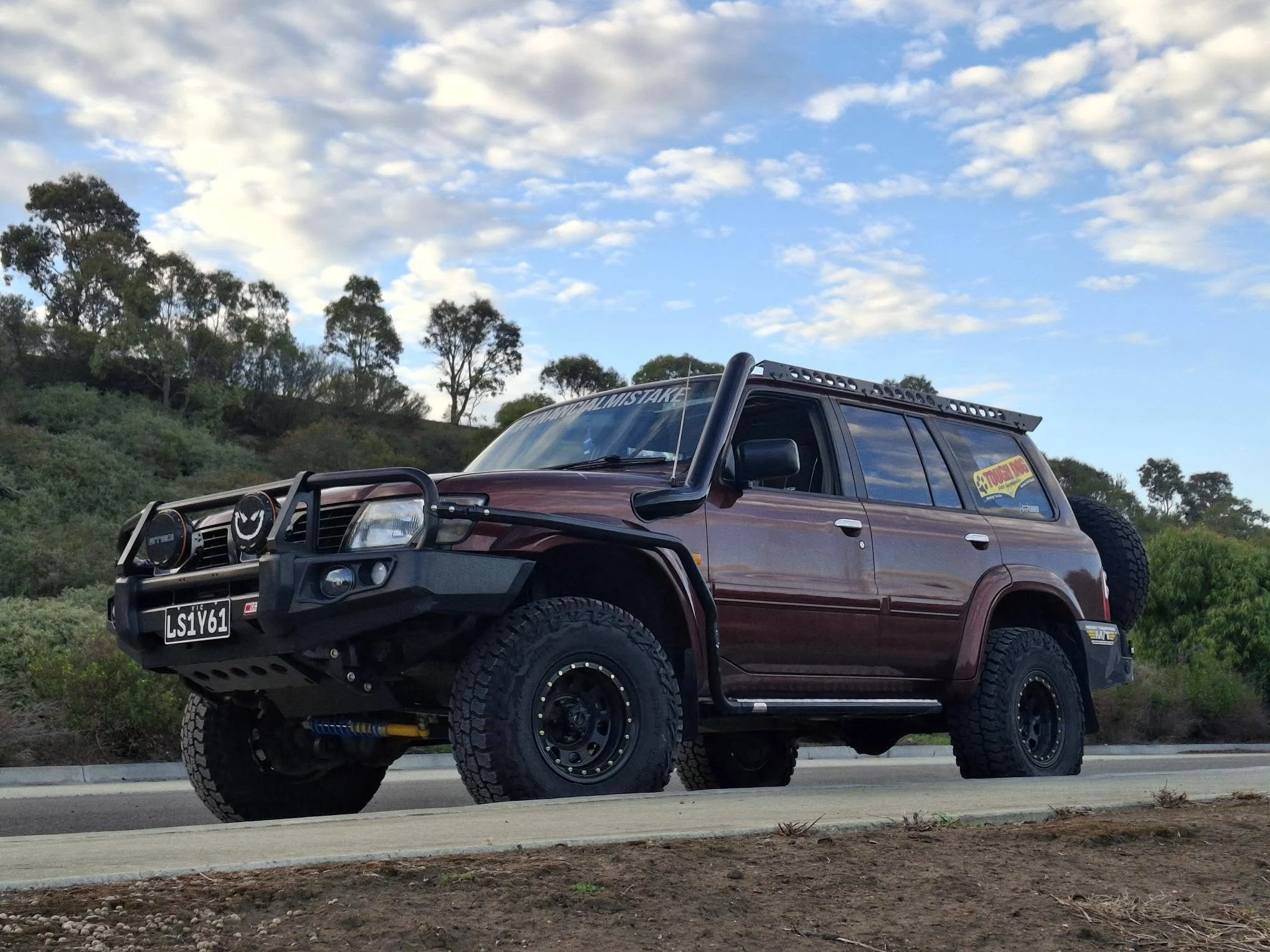 A red off-road vehicle parked on a roadside with trees and a partly cloudy sky in the background.