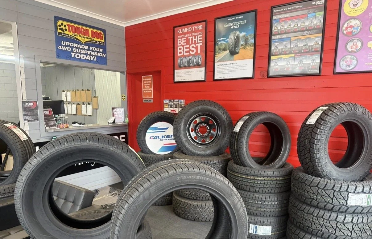 A tire shop with stacked tires and tire posters on the red wall, including one for Falken tires and promotional posters for Kumho tires and car brands. There is a small counter area in the background with signs and a window.