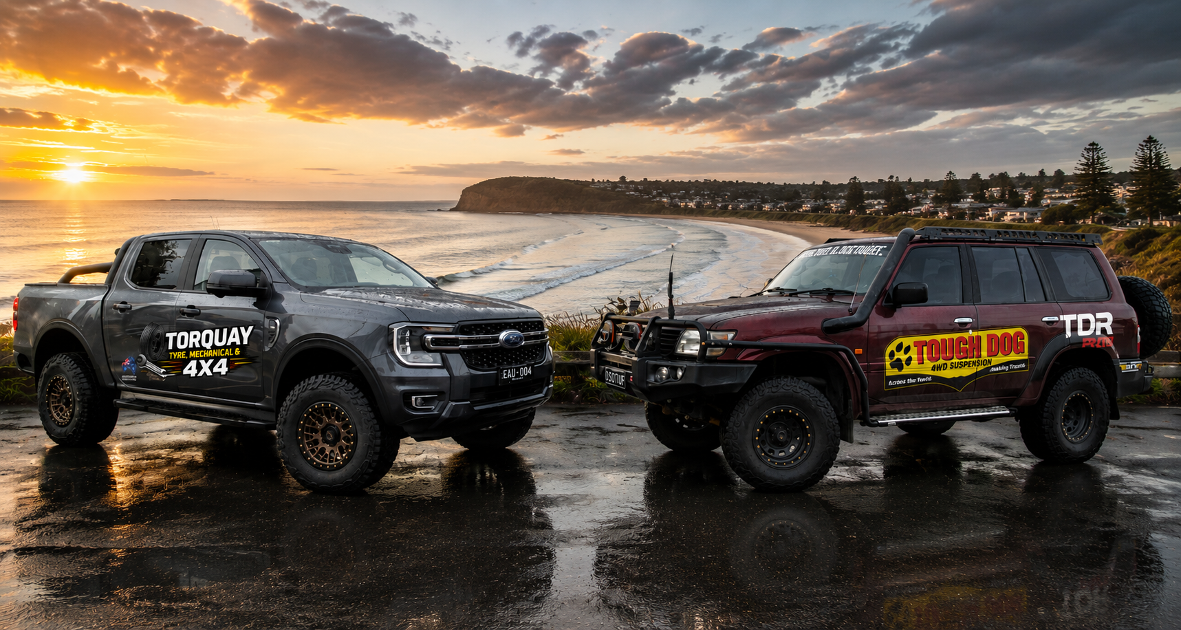 Two off-road vehicles parked on a wet road near a coastal view at sunset, with clouds, ocean waves, and distant houses in the background.