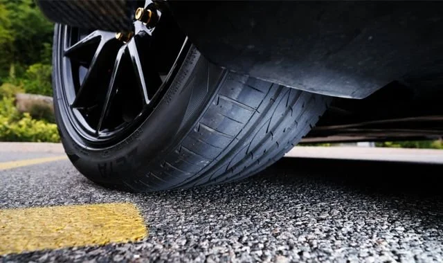 Close-up of a car tire on a paved road, showing black alloy rim and yellow lane line.