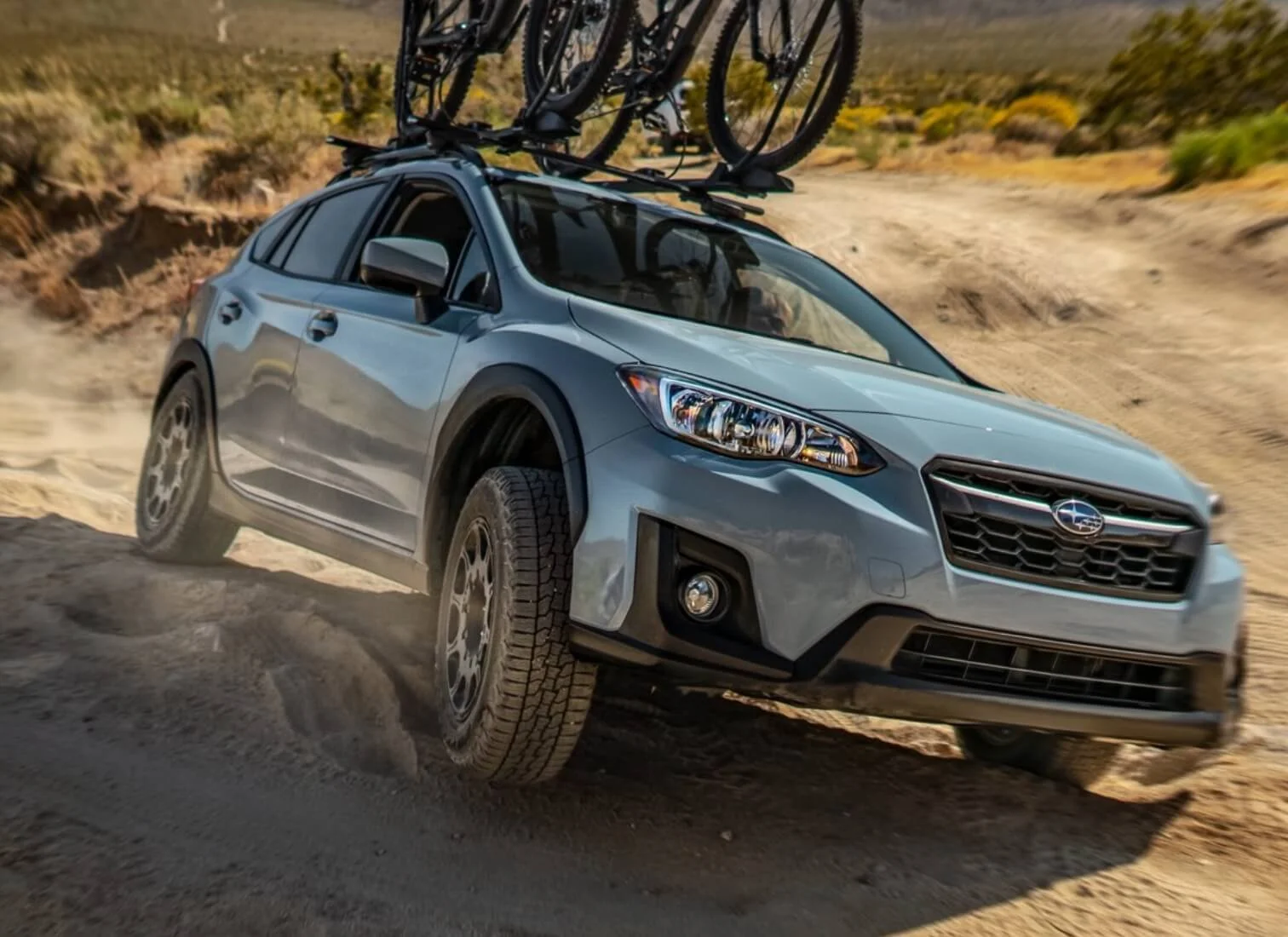 A silver Subaru crossover SUV driving on a dirt trail in a desert landscape, with bicycle racks carrying bikes on the roof.