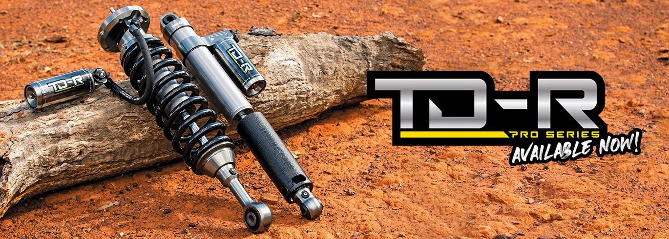 A pair of shock absorbers laying on dirt next to a large piece of wood, with a graphic text overlay advertising T-R Pro Series shock absorbers, available now.