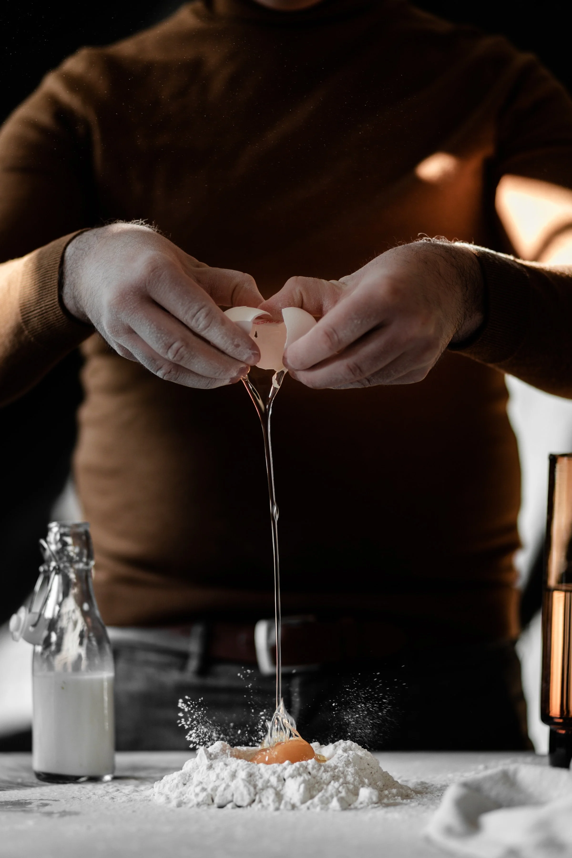 Person cracking an egg into a pile of flour with a milk bottle nearby, on a black table with a blurred background.