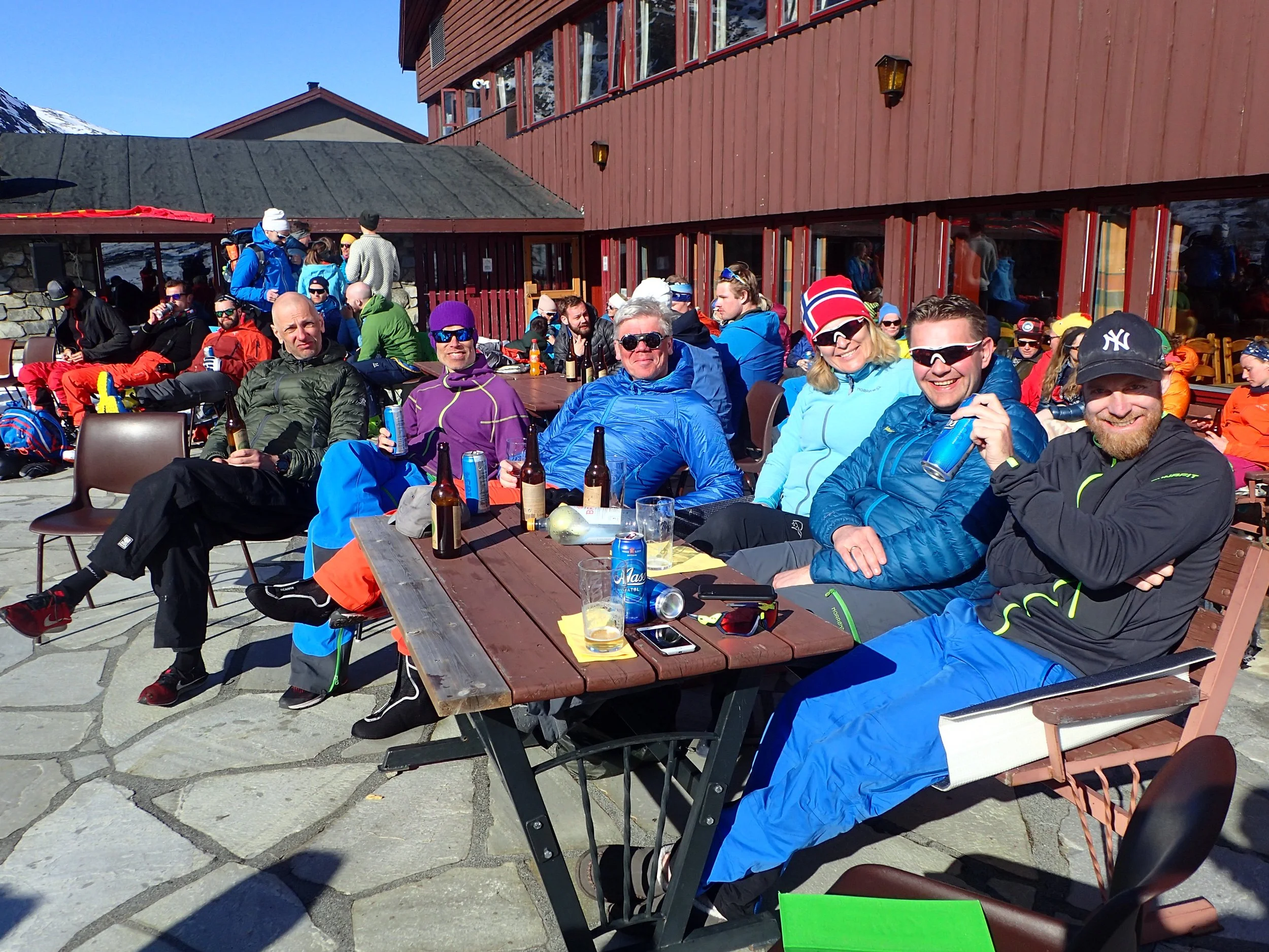 En gruppe mennesker sitter på utekafé med fjell i bakgrunnen. De er kledd i ski- og fjellklær, og mange har drikke i hendene. Det er sol og flere har på solbriller.